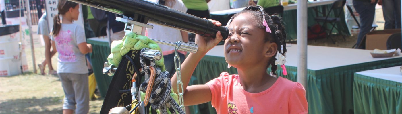Young African American girl looking through telescope while at a 4-H booth at a fair.