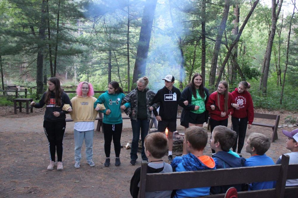 Eight camp counselors stand in a line with arms linked, ready to perform for seated campers