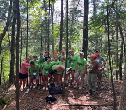 A dozen people, mostly teenagers and adults, stand on top of a hill covered by trees. 